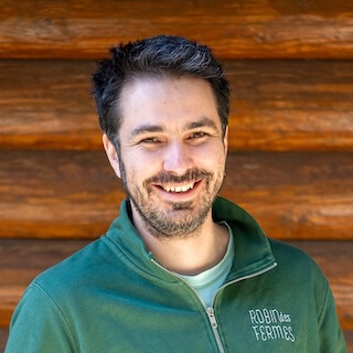 Portrait de Fabien portant un t-shirt rayé blanc et bleu, bras croisés, avec un fond de verdure.
